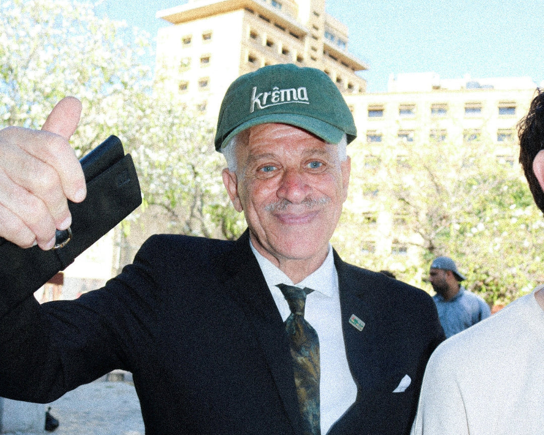 Man wearing a 'Krema' cap and suit, giving a thumbs-up outdoors with buildings and trees in the background.